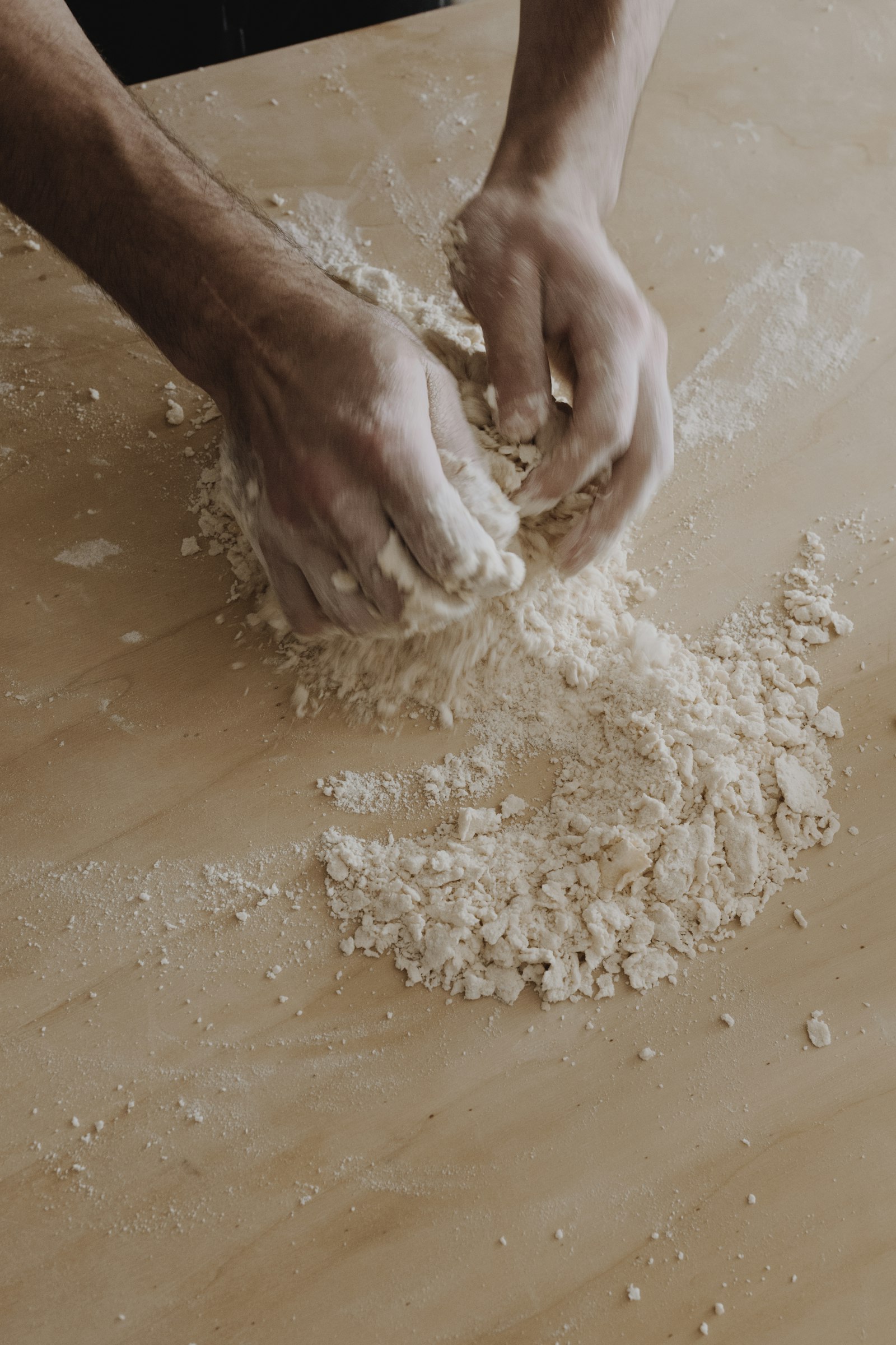 A baker's hands working flour on a pale wooden work surface