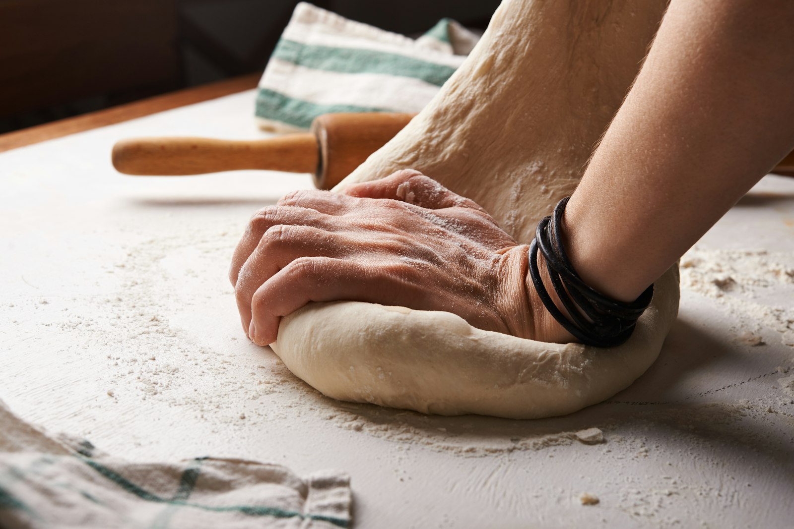 Hands kneading sourdough on a floured work surface beside a rolling pin and linen cloth